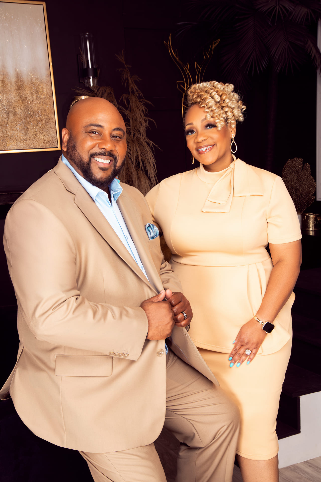 A man in a beige suit and a woman in a cream dress posing together indoors.