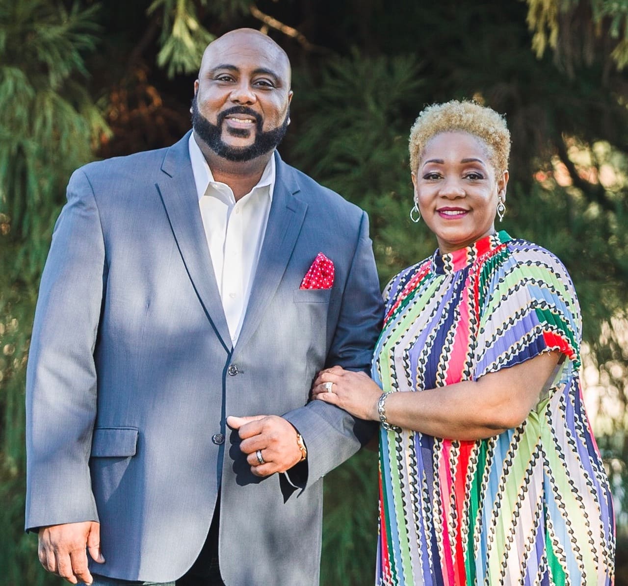 A well-dressed couple smiling outdoors with greenery in the background.